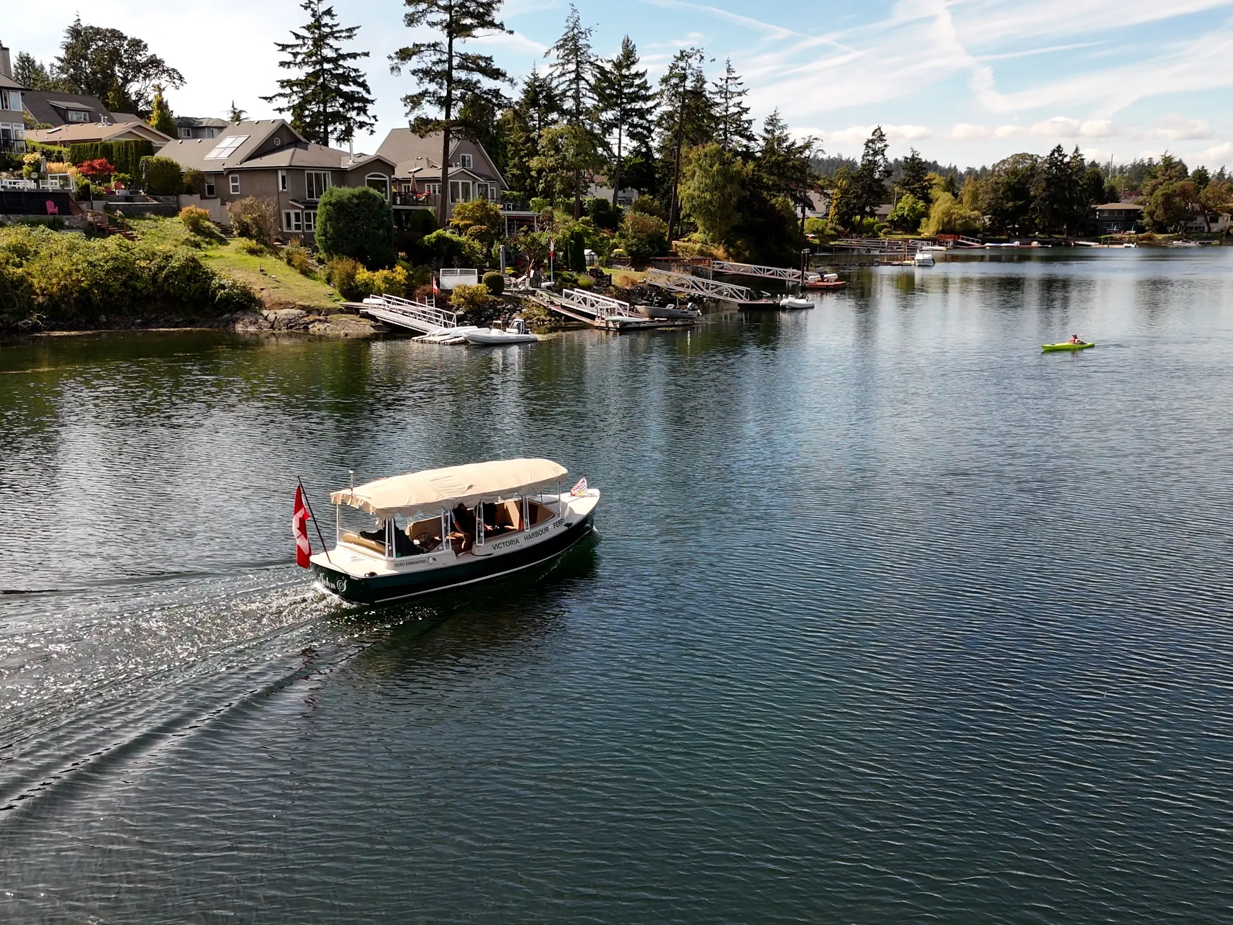 A small boat with a canopy sails near a shoreline with houses and trees.
