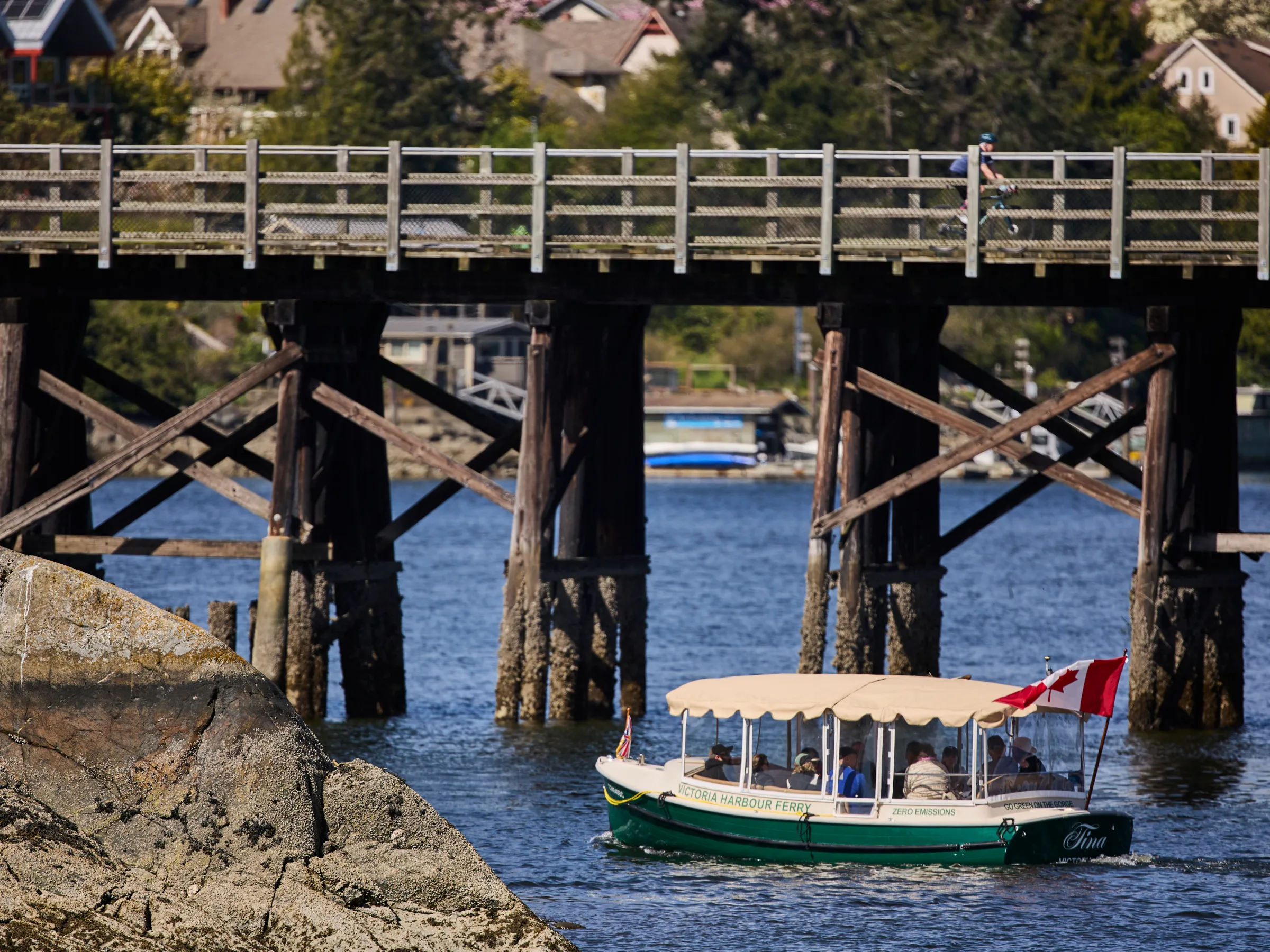 Boat with canopy under wooden bridge on calm water, with rocky foreground.