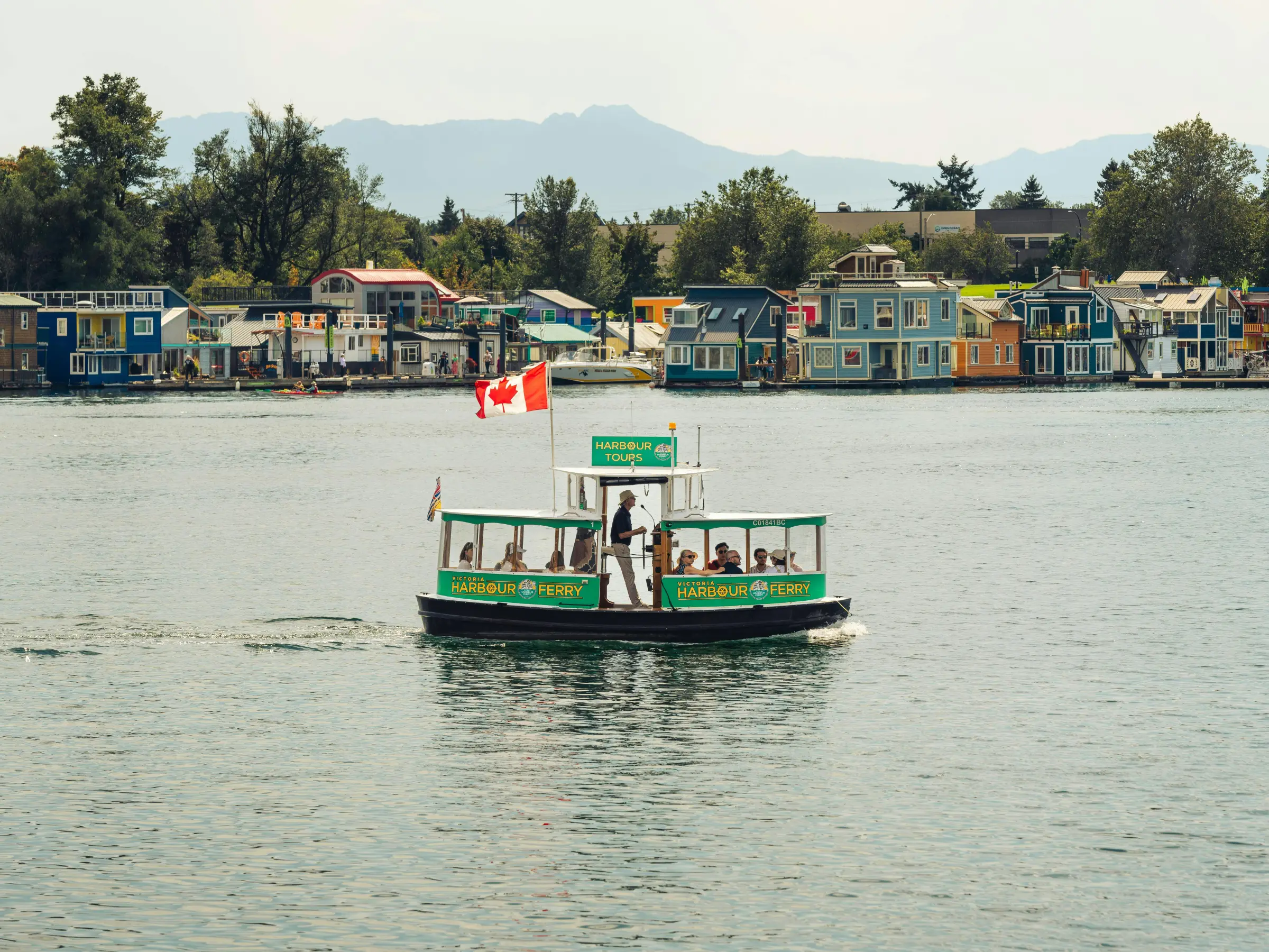 Small ferry with Canadian flag on water near colorful houseboats.