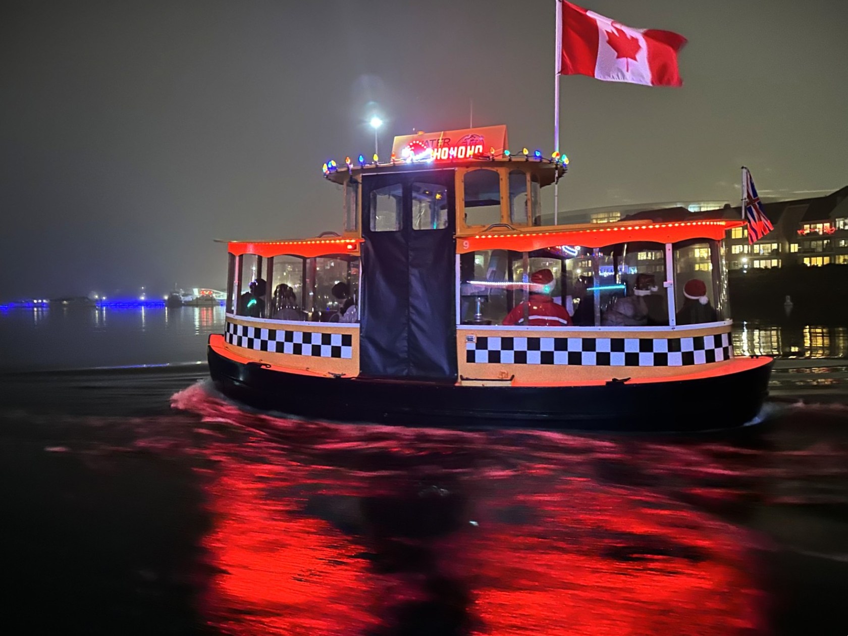 A small boat with a Canadian flag, decorated with lights, reflects on water at night.