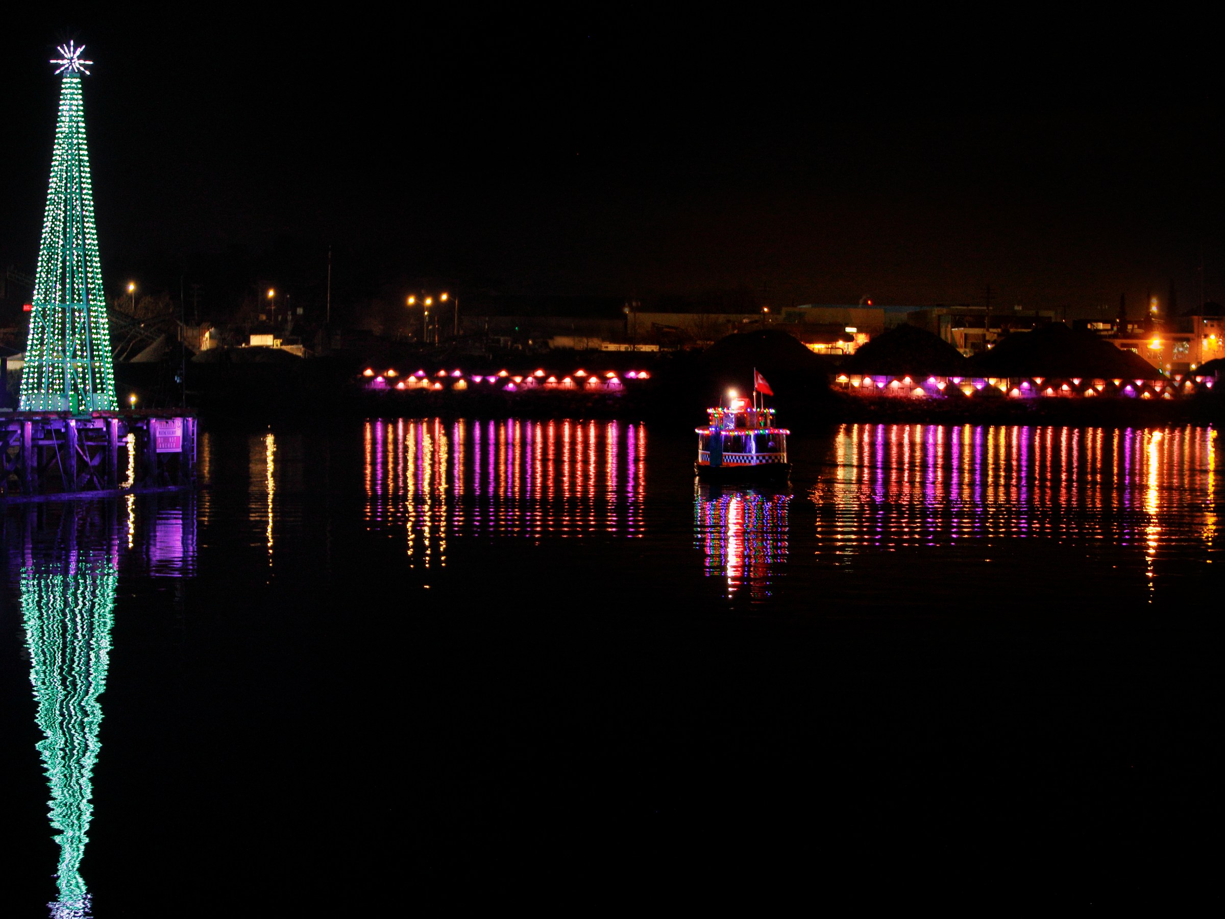 Lit Christmas tree and decorated boat reflected in water at night.