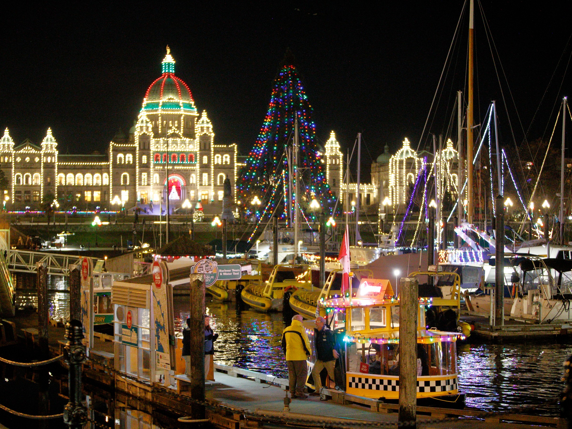 Night harbor scene with festive lights on buildings and boats, featuring a lit dome and Christmas tree.