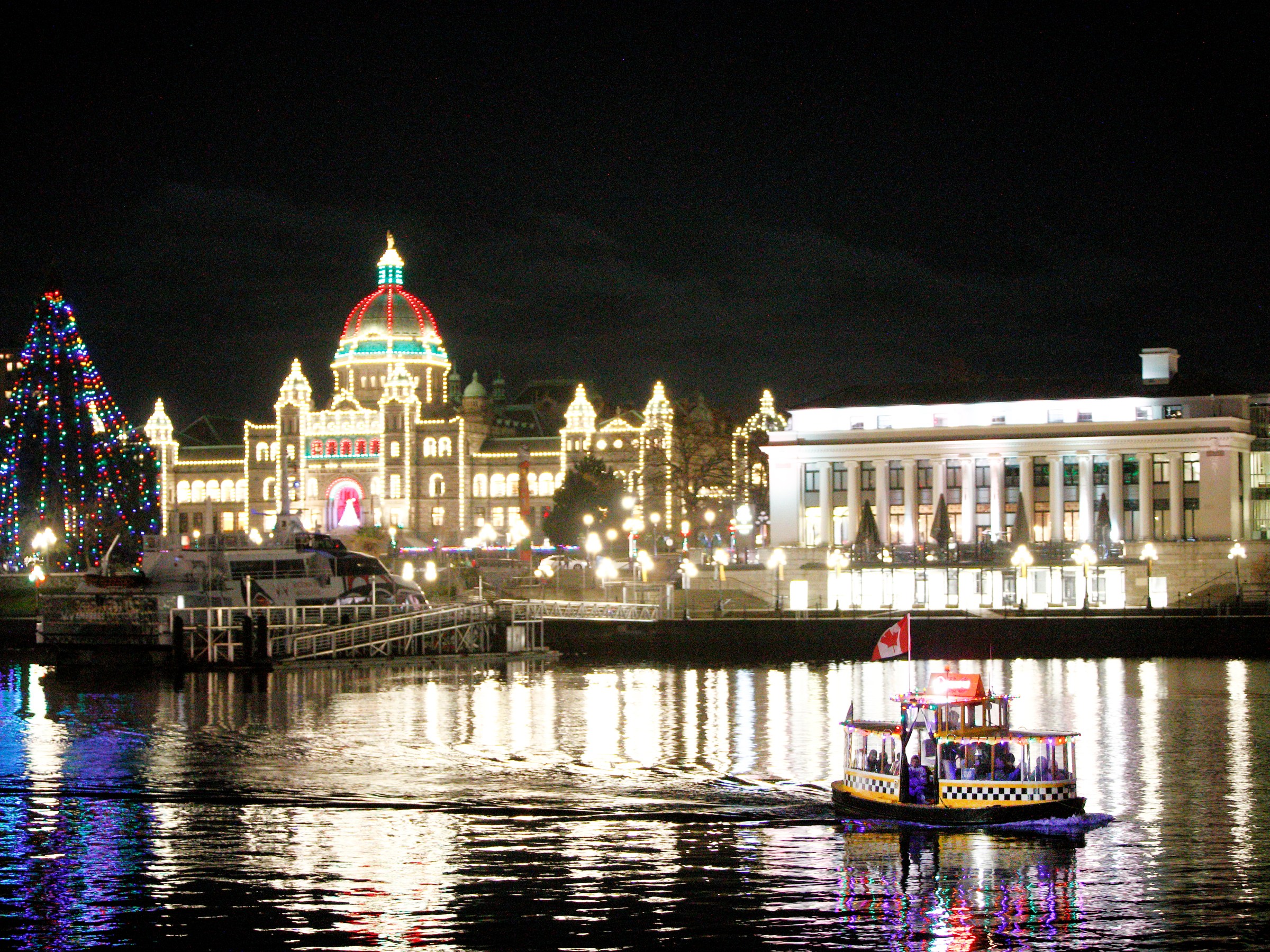 Night view of a lit building and decorated tree reflecting in water, with a small ferry in the foreground.