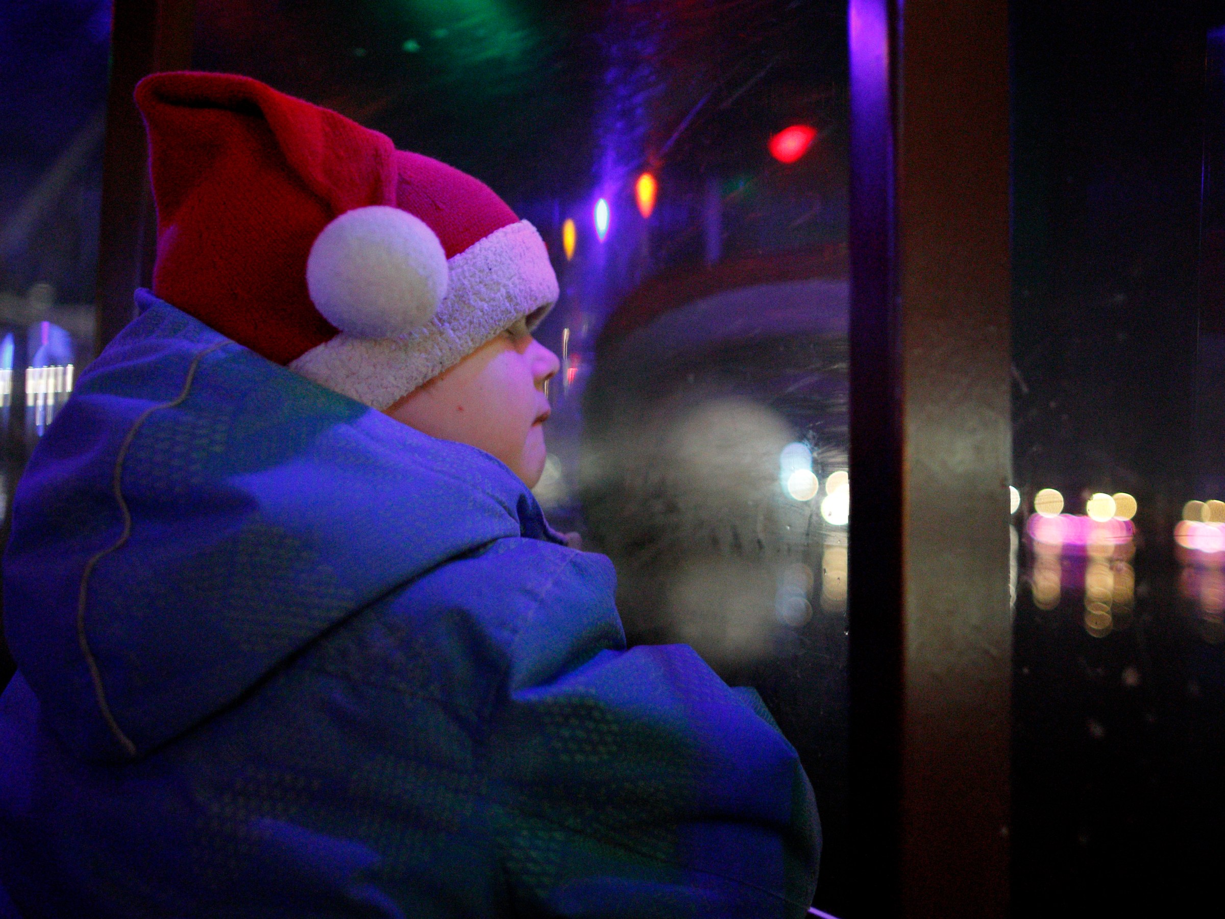 Child in Santa hat looks out window at night with colorful lights reflected on glass.