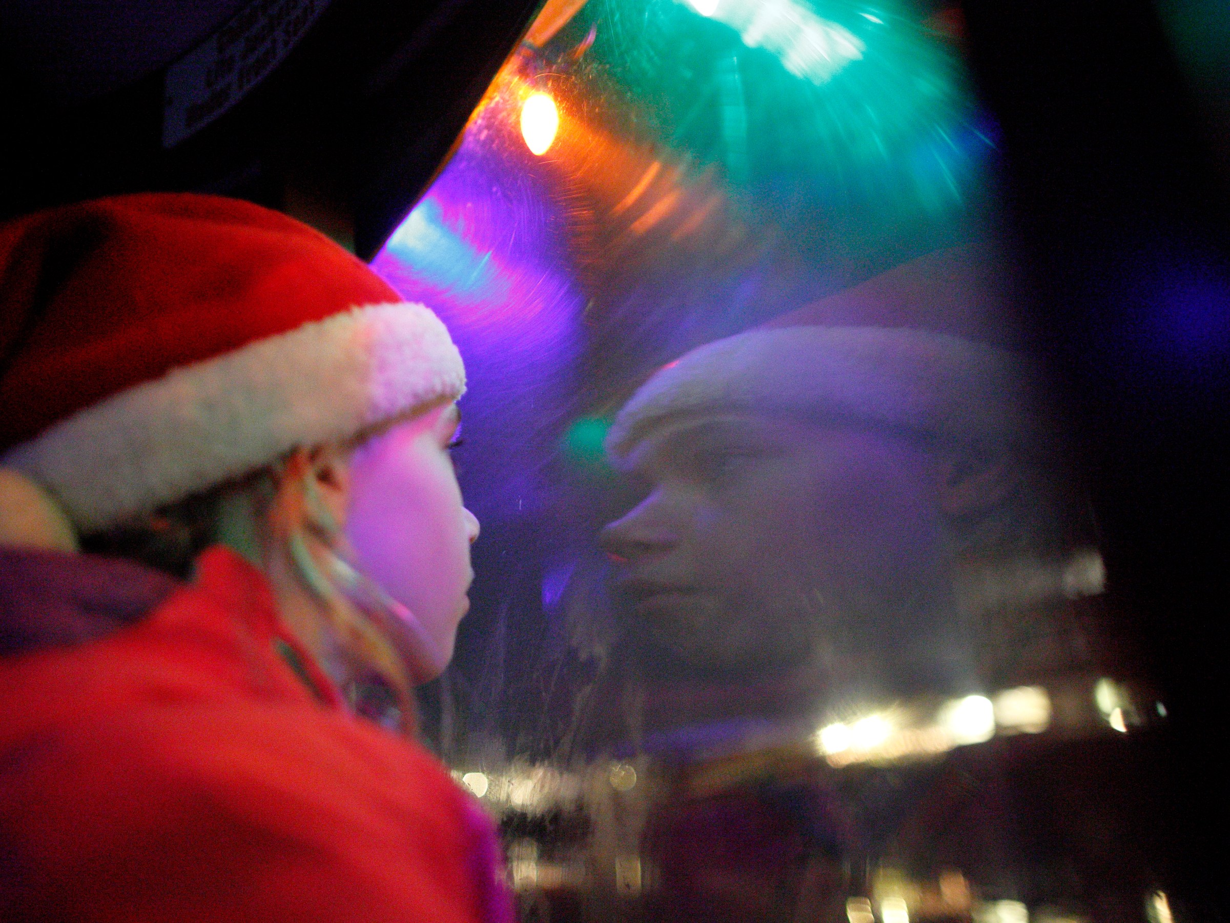 Child in Santa hat looking at colorful lights through a window.